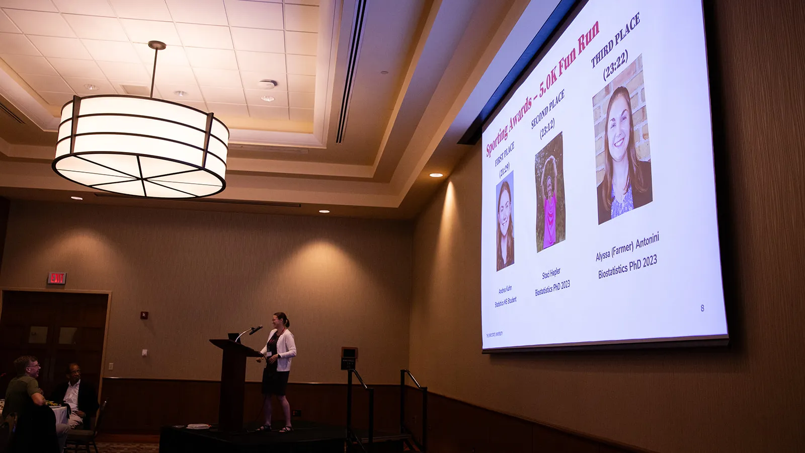 Elly Kaizar standing at a podium with a screen presentation of the award winners
